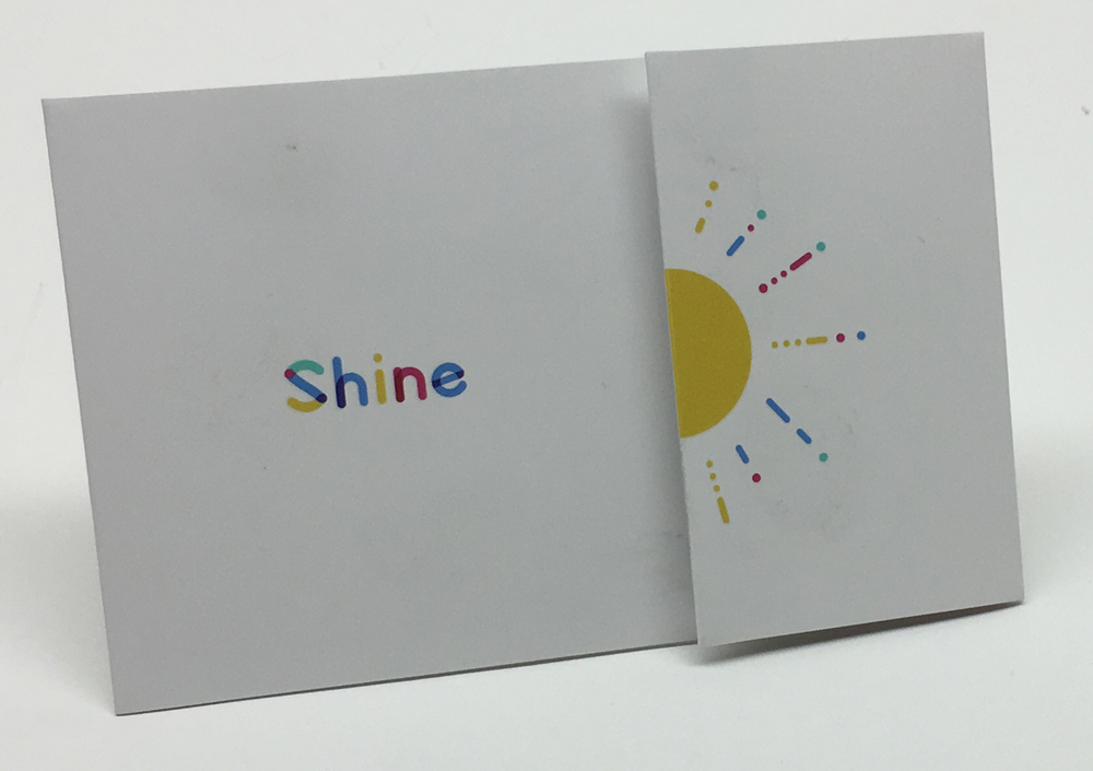 Carte de rangement ou pochette double avec le mot “Shine” coloré et un soleil stylisé sur fond clair.