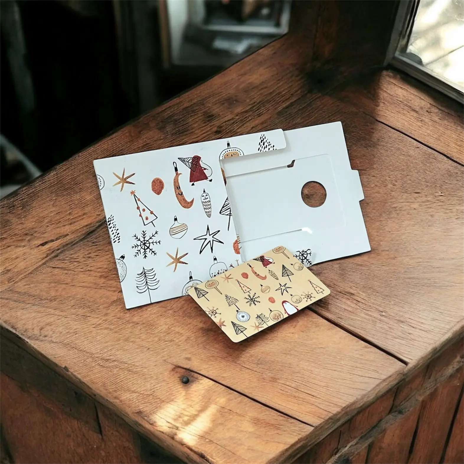 Pochettes et carte décorative décorées de motifs Noël sur papier kraft et blanc, posées sur une table en bois.